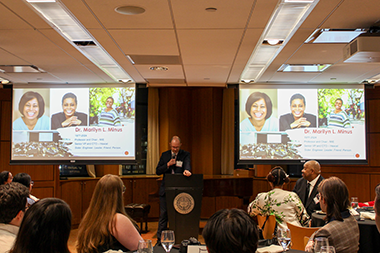 Andrew Gouldstone speaking at a podium between two projections of Marilyn Minus